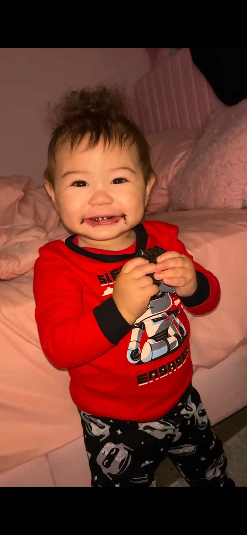 Adonis is registered to the contest to win money with this photo: toddler, child, smiling, chocolate, cookie, pajamas, red_shirt, robot_design, bedroom, bed, blanket, pink, happy, indoor, person, cute, hair_up, holding, standing, face