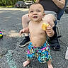 asphalt, baby_bottle, bare_chest, car, chalk_drawing, child, colorful, daylight, food, grass, leg_brace, outdoor, person, road, shoe, shorts, sitting, smiling, toddler, toy