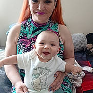 Raeleigh is registered to the contest to win money with this photo: baby, child, woman, smile, happy, indoor, clothing, sitting, person, face, hair, makeup, holding, baby_clothes, patterned_dress, furniture, blanket, home, portrait, cute