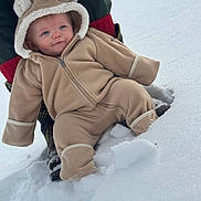 Alexander is registered to the contest to win money with this photo: baby, child, snow, winter_clothing, snowsuit, hood, face, outdoor, cold_weather, fence, boots, red_socks, person, candid, cute, smile, infant, warm_clothing, fuzzy, support