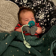 Atticus is registered to the contest to win money with this photo: baby, pacifier, mittens, blanket, star_pattern, moon_pattern, sleeping, infant, cozy, red_striped_clothing, face, head, child, indoor, comfort, cute, resting, young, human, portrait