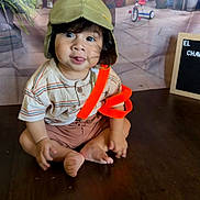 Luca is registered to the contest to win money with this photo: baby, backdrop, child, curly_hair, cute, hat, indoor, person, pilot_hat, playful, portrait, shorts, signboard, sitting, smiling, striped_shirt, toddler, tongue_out, tricycle, wooden_floor