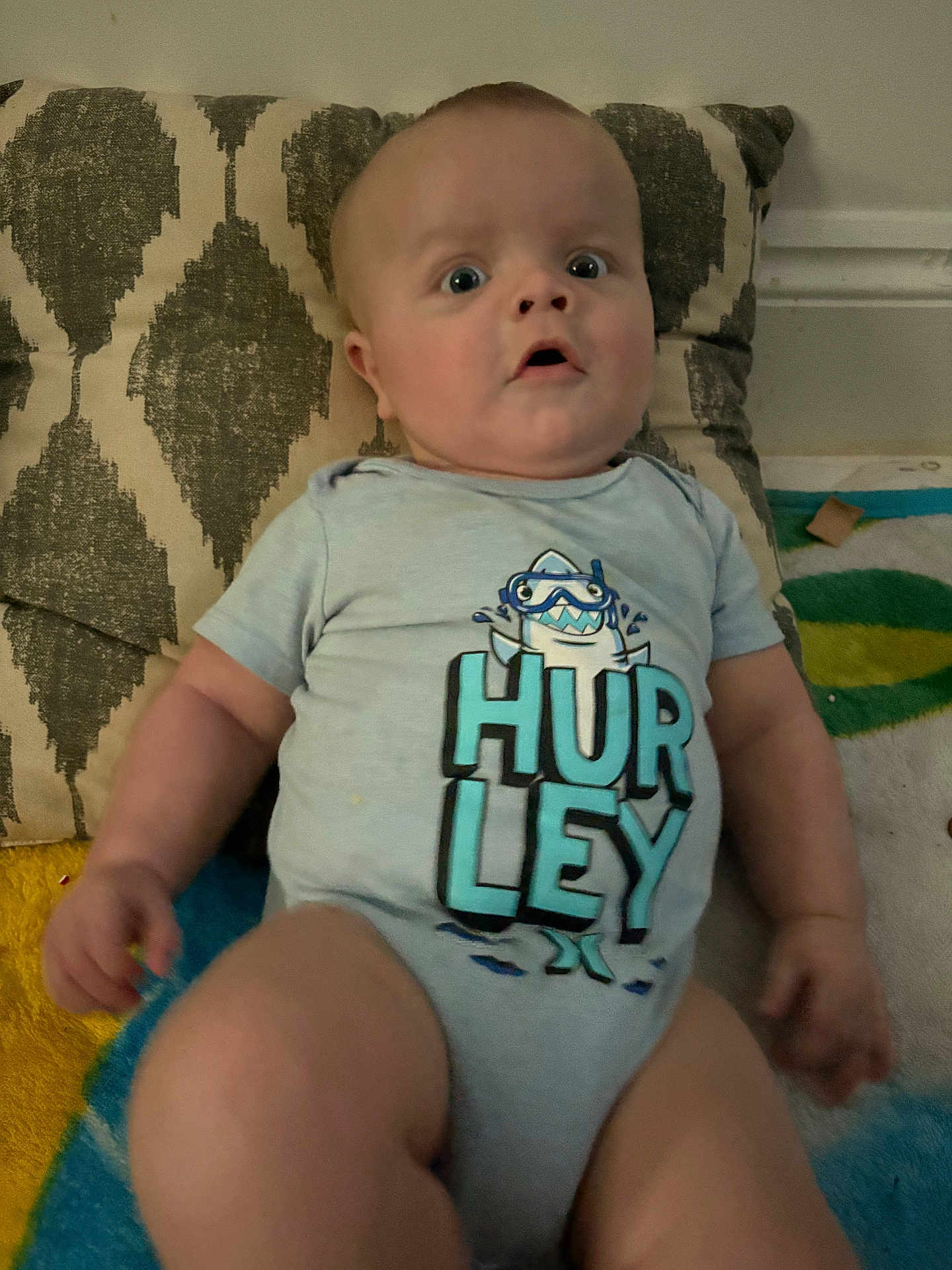 Andrew joined the competition — help win amazing prizes! baby, infant, child, onesie, clothing, face, expression, pillow, blanket, pattern, indoor, person, cute, wide_eyes, mouth_open, sitting, soft_toys, home, floor, colorful