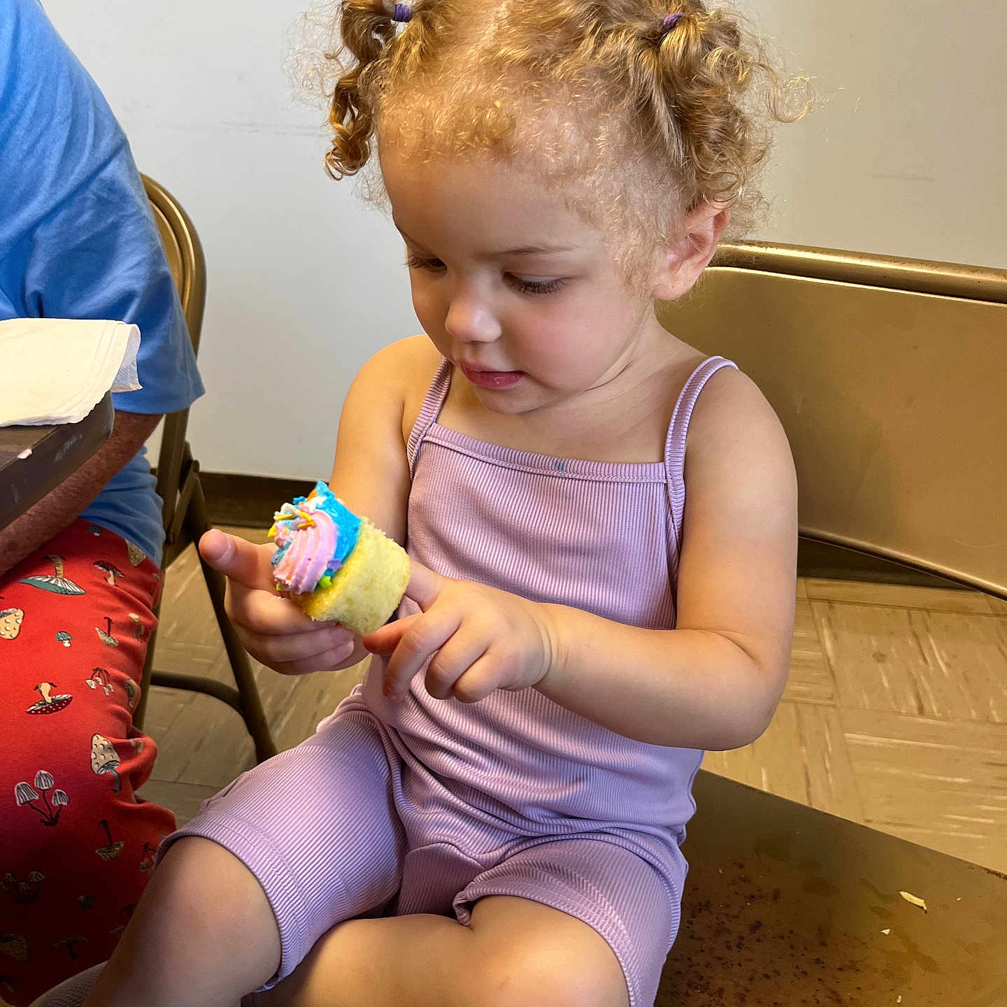 Lillyannah is registered to the contest to win money with this photo: adult, casual, chair, child, cupcake, curly_hair, cute, fingers, floor, food, frosting, hair_ties, indoor, person, playful, purple_clothing, sitting, snack, table, toddler