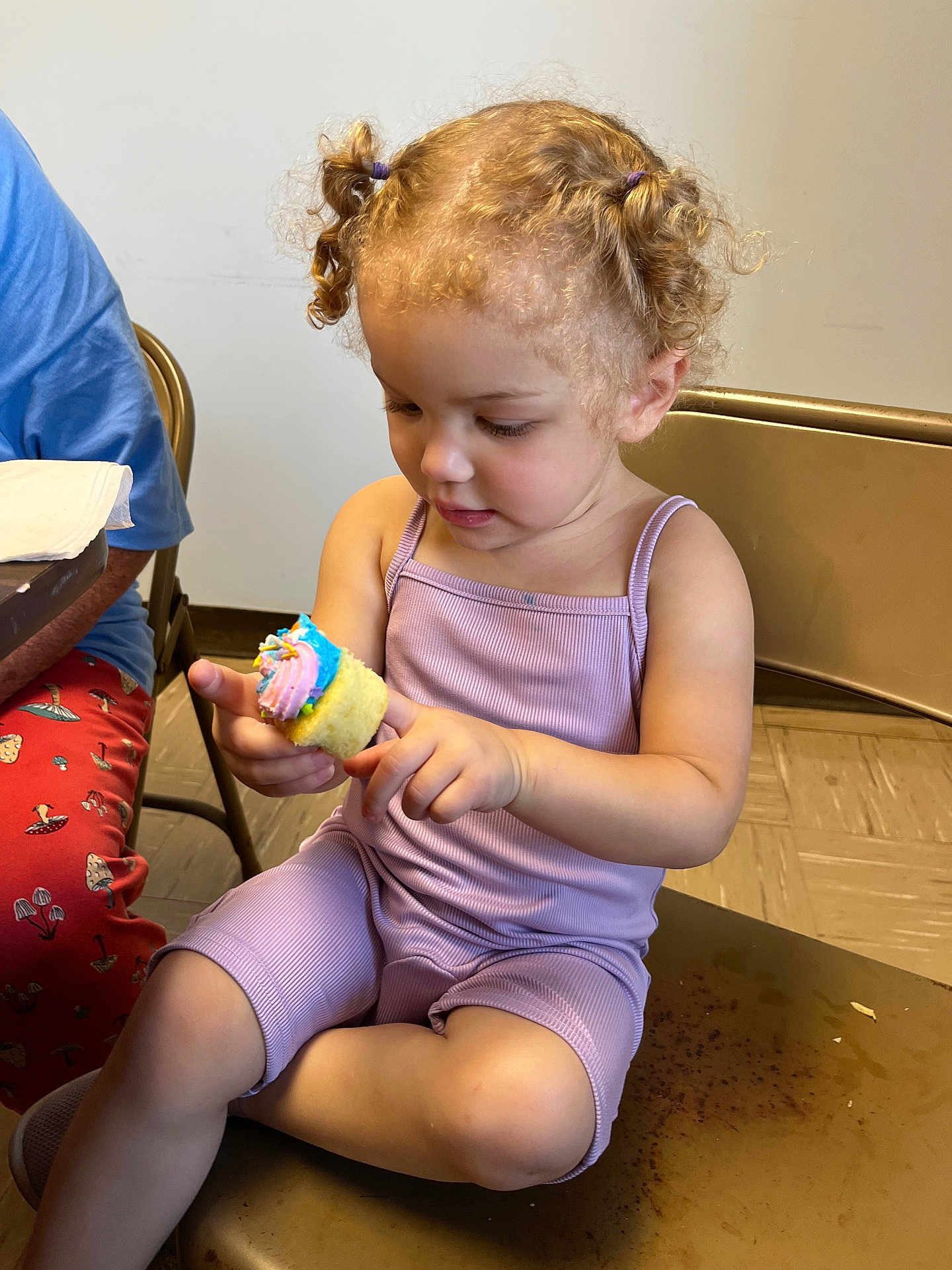 Lillyannah is registered to the contest to win money with this photo: toddler, child, cupcake, food, purple_clothing, chair, curly_hair, indoor, person, sitting, table, adult, floor, snack, playful, cute, hair_ties, frosting, fingers, casual