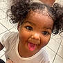 child, toddler, curly_hair, pigtails, tongue_out, smiling, white_shirt, flower_design, indoor, tile_floor, cute, playful, portrait, person, happy, young_child, closeup, funny_face, girl, casual_clothing