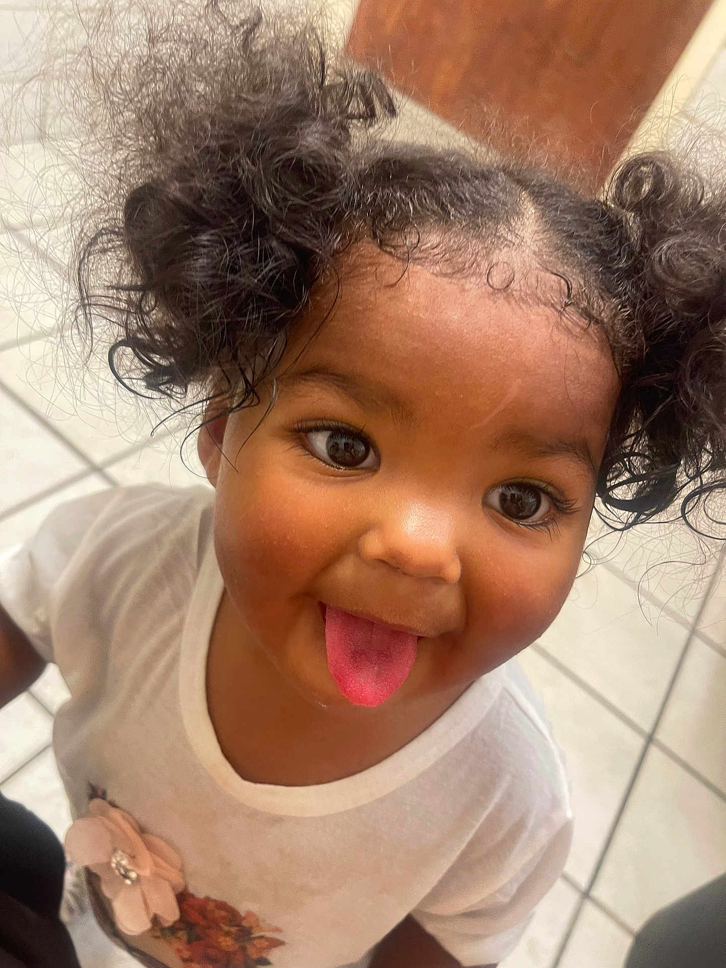 Z'Naya joined the competition — help win amazing prizes! child, toddler, curly_hair, pigtails, tongue_out, smiling, white_shirt, flower_design, indoor, tile_floor, cute, playful, portrait, person, happy, young_child, closeup, funny_face, girl, casual_clothing