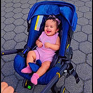 Sama is registered to the contest to win money with this photo: baby, child, stroller, smiling, laughing, outdoor, sidewalk, pavement, blue_stroller, pink_clothing, pink_socks, curly_hair, sitting, infant, portrait, hand, pavement_pattern, park, daylight, happy