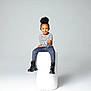 child, smiling, portrait, studio, minimalist, black_bow, striped_shirt, blue_jeans, black_boots, cylindrical_pedestal, high_puff_hairstyle, seated, fashion, happy, casual_clothing, young_kid, isolated_background, cute, modern, confident
