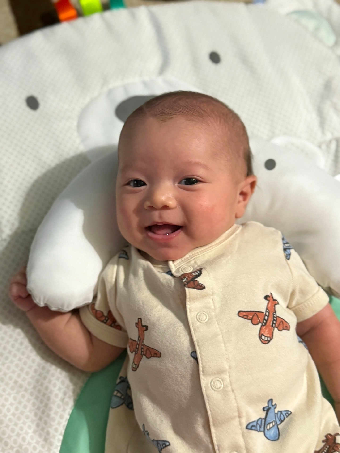 Ashton is registered to the contest to win money with this photo: baby, smiling, infant, onesie, airplane_pattern, cushion, bear_shape, soft_toy, cute, happy, child, portrait, lying_down, indoors, closeup, face, head, baby_clothing, fabric, comfort