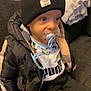 Kassim Menassel a rejoint le concours — aidez-le/la à gagner de superbes lots ! baby, beanie, black_jacket, casual, child, clothing, couch, curious, face, footwear, hand, headwear, indoor, pacifier, pants, person, puffer_jacket, puma, sitting, young