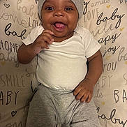 Kannon is registered to the contest to win money with this photo: baby, beanie, child, closeup, comfortable, cozy, cushion, cute, gray_pants, happy, indoor, infant, lying_down, person, playful, portrait, smiling, text_pattern, tongue_out, white_shirt