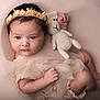 baby, infant, flower_headband, knitted_toy, teddy_bear, cream_dress, soft_lighting, portrait, cute, cozy, cushion, newborn, child, adorable, resting, soft_texture, innocence, closeup, indoor, peaceful