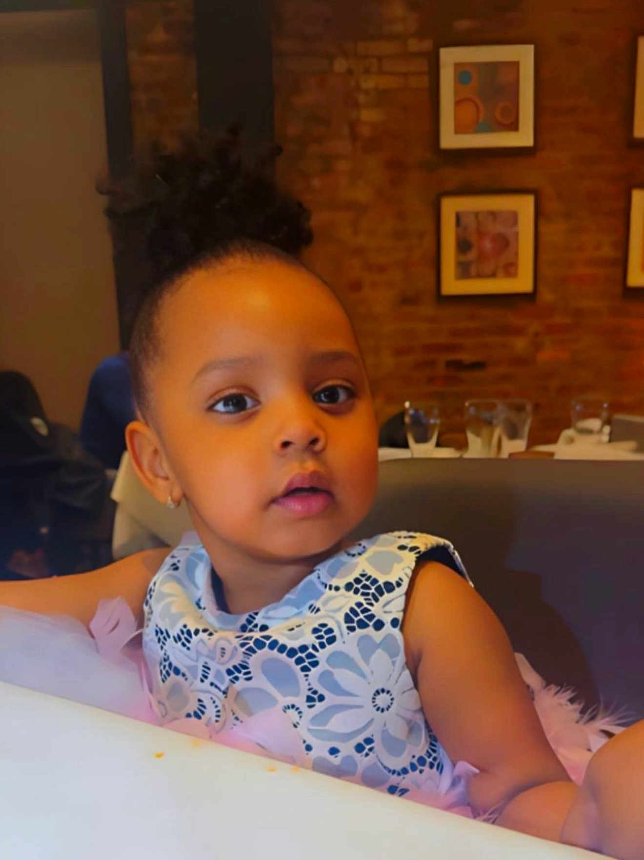 Cherifah joined the competition — help win amazing prizes! artwork, brick_wall, child, curly_hair, cute, dress, earrings, face, girl, glass, hair, indoor, looking_away, patterned_clothing, portrait, restaurant, seated, skin, table, toddler