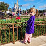 child, girl, purple_shirt, pink_pants, fence, greenery, castle, theme_park, outdoor, daylight, people, blue_sky, clouds, tourists, walking, concrete, shoes, hairstyle, sunny, scenic