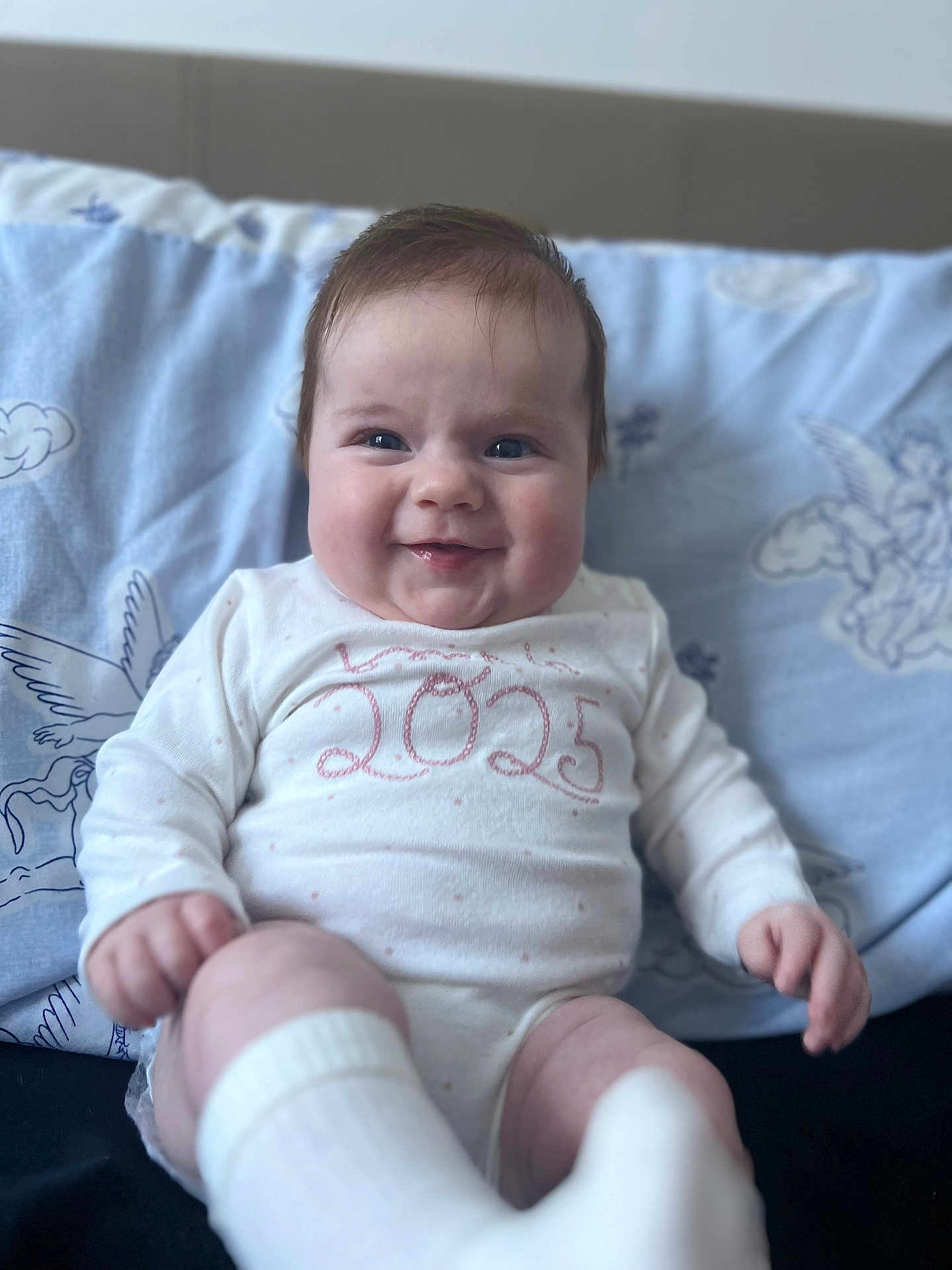 Delilah-Mae is registered to the contest to win money with this photo: baby, infant, smiling, onesie, white_clothing, socks, chubby_cheeks, face, person, indoor, blanket, patterned_blanket, blue_background, happy, cute, child, soft_lighting, portrait, seated, young