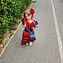 child, girl, dress, red, black, flower, hair_accessory, playful, outdoor, path, greenery, bushes, pavement, footwear, posing, young, fashion, accessory, smile, casual