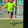 child, running, green_shirt, shorts, playground, water_spray, mural, flower_mural, grass, outdoor, happy, smiling, sunny, play_area, splash_pad, colorful, fun, active, person, summer