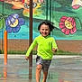 child, running, splash_pad, water, green_shoes, neon_shirt, shorts, smiling, outdoor, mural, wall_painting, flowers, cats, playground, summer, fun, happy, motion, water_splash, daytime