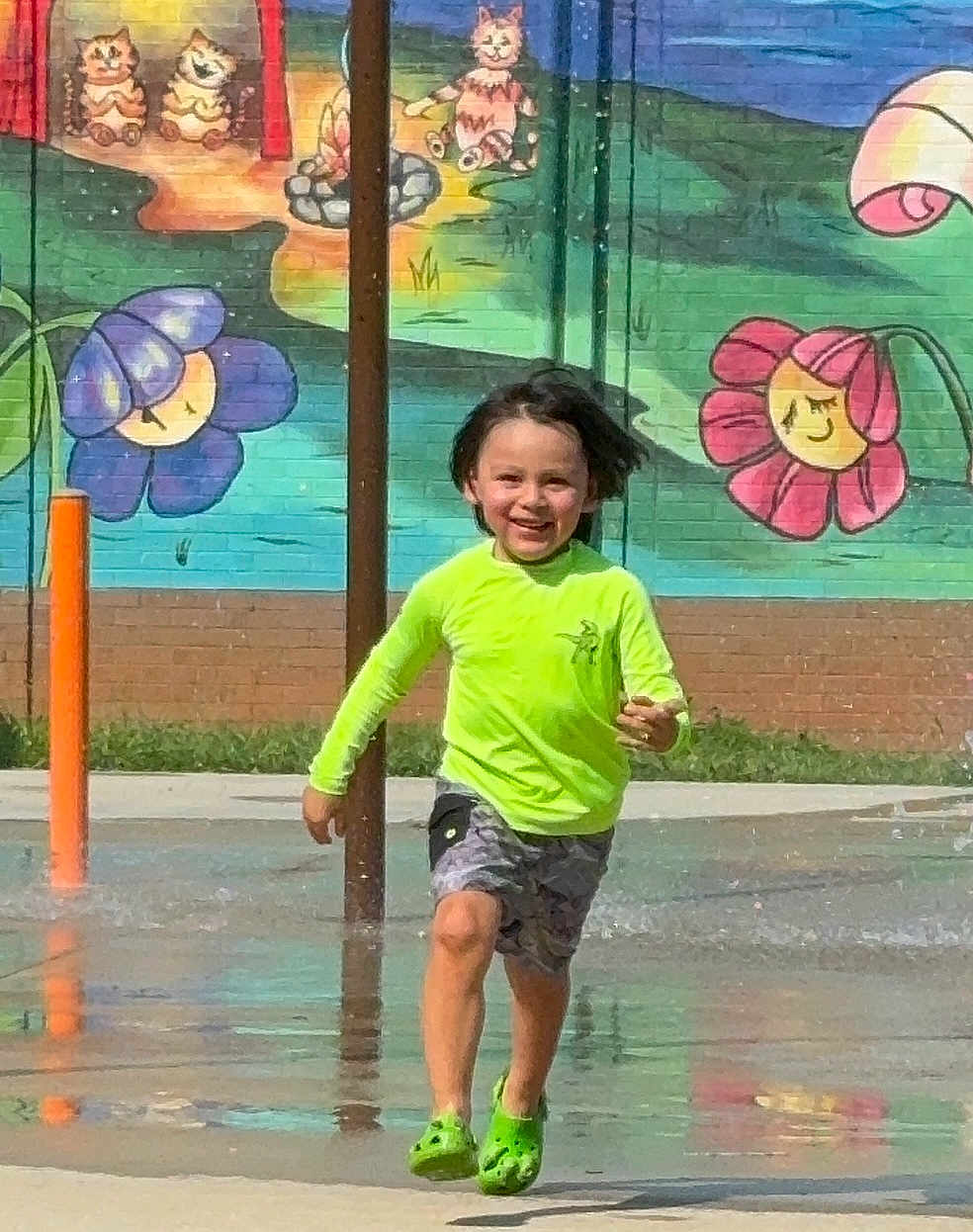 Elias joined the competition — help win amazing prizes! child, running, splash_pad, water, green_shoes, neon_shirt, shorts, smiling, outdoor, mural, wall_painting, flowers, cats, playground, summer, fun, happy, motion, water_splash, daytime
