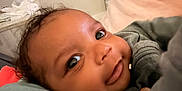 Nasir is registered to the contest to win money with this photo: baby, face, close_up, smile, eyes, infant, lying_down, cushion, blanket, indoors, child, soft_texture, cozy, curly_hair, expression, person, portrait, young, cute, warm