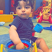 Kaïs Habeche participe au concours pour gagner de l'argent avec cette photo : toddler, child, curly_hair, striped_shirt, indoor, playroom, toys, colorful, buckets, plastic_toys, floor, footwearless, seated, expression, curious, hand, blue_cup, yellow_handle, play, daylight