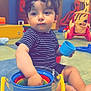 toddler, child, curly_hair, striped_shirt, indoor, playroom, toys, colorful, buckets, plastic_toys, floor, footwearless, seated, expression, curious, hand, blue_cup, yellow_handle, play, daylight