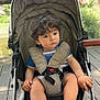 child, toddler, stroller, outdoor, curly_hair, blue_shirt, sandals, seatbelt, wooden_deck, trees, building, daylight, person, baby, nature, park, hand, face, expression, sunlight