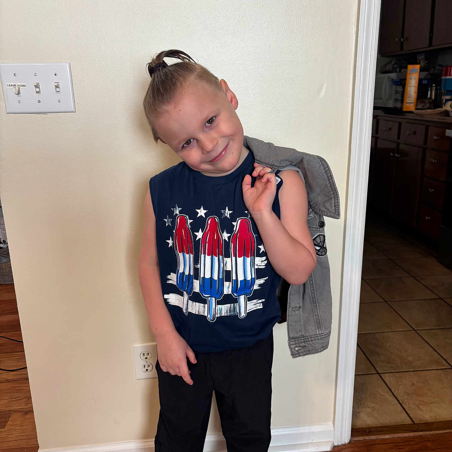 Carson is registered to the contest to win money with this photo: child, boy, indoor, pose, smile, casual_clothing, sleeveless_shirt, popsicle_print, jacket, hair_style, wooden_floor, wall, light_switch, electrical_outlet, kitchen, cabinet, tile_floor, doorway, standing, happy