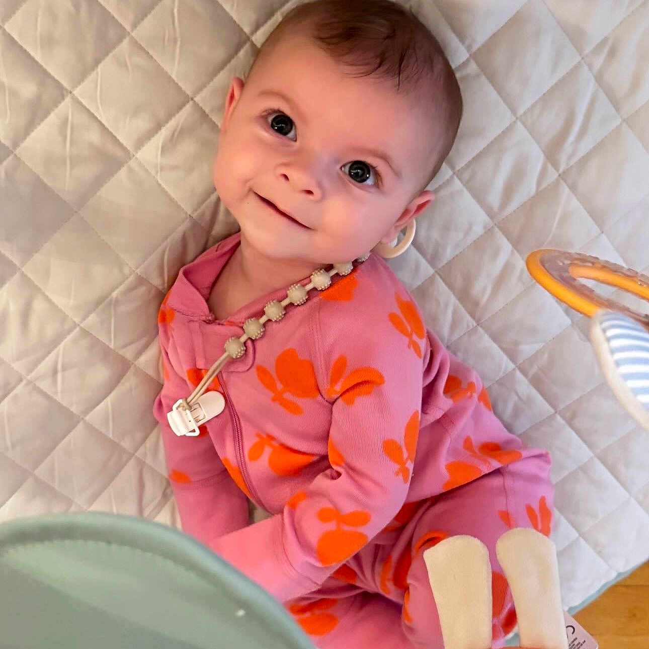 Calliope is registered to the contest to win money with this photo: baby, child, smiling, pink_clothing, orange_flower_pattern, onesie, quilted_mat, toy, indoor, floor, cute, happy, infant, person, portrait, playtime, soft_toys, head, eyes, skin