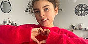 Alaïs participe au concours pour gagner de l'argent avec cette photo : child, girl, red_sweater, heart_hands, indoor, living_room, clock, mirror, furniture, smiling, face, person, cozy, decor, hair, earrings, sleeves, casual_clothing, young, portrait