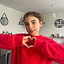 child, girl, red_sweater, heart_hands, indoor, living_room, clock, mirror, furniture, smiling, face, person, cozy, decor, hair, earrings, sleeves, casual_clothing, young, portrait