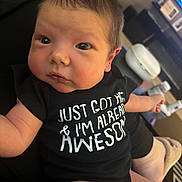 Brayden is registered to the contest to win money with this photo: baby, infant, onesie, black_clothing, face, eyes, arm, hand, indoor, living_room, television, furniture, person, cute, newborn, skin, expression, sitting, home, relaxed