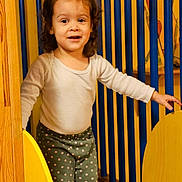 Jade a rejoint le concours — aidez-le/la à gagner de superbes lots ! child, toddler, curly_hair, play_area, yellow, blue, wooden_structure, long_sleeve_shirt, polka_dot_pants, indoor, smiling, standing, person, clothing, happy, kid, cute, playground, fun, bright_colors