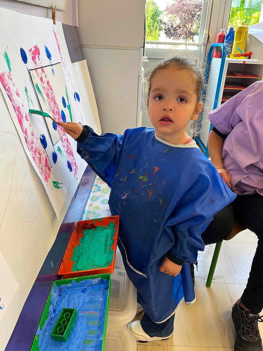 Maëlle a rejoint le concours — aidez-le/la à gagner de superbes lots ! child, painting, art, smock, paintbrush, easel, paint_tray, green_paint, blue_paint, indoor, floor, window, adult, person, artwork, creative, colorful, clothing, footwear, expression