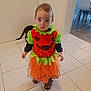 baby, black_cat, cat, child, costume, curious, cute, dress, floor, green, halloween, holiday, indoor, orange, person, pumpkin, shoes, standing, tile_floor, toddler