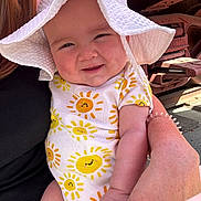 Carlisa is registered to the contest to win money with this photo: baby, child, smiling, sun_hat, onesie, sun_pattern, adult_hand, outdoor, sunlight, cute, happy, face, arm, person, holding, red_metal, background, warm, skin, infant