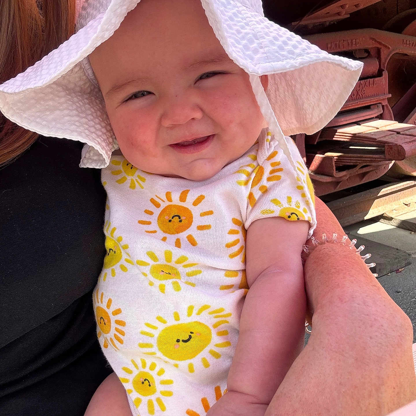 Carlisa is registered to the contest to win money with this photo: baby, child, smiling, sun_hat, onesie, sun_pattern, adult_hand, outdoor, sunlight, cute, happy, face, arm, person, holding, red_metal, background, warm, skin, infant