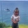 Tania a rejoint le concours — aidez-le/la à gagner de superbes lots ! child, girl, smiling, plush_toy, white_tshirt, denim_shorts, sneakers, grass, glass_railing, ocean, rocky_islet, cloudy_sky, outdoor, standing, happy, casual_clothing, topknot_hairstyle, nature, coast, scenic_view