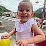child, toddler, smiling, outdoor, table, yellow_object, clear_object, white_clothing, short_hair, plastic, red_chair, houses, greenery, mountains, cloudy_sky, decorative_hats, wires, street, happy, play