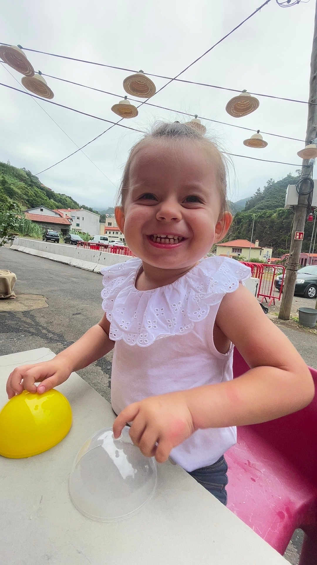 Tania participe au concours pour gagner de l'argent avec cette photo : child, toddler, smiling, outdoor, table, yellow_object, clear_object, white_clothing, short_hair, plastic, red_chair, houses, greenery, mountains, cloudy_sky, decorative_hats, wires, street, happy, play