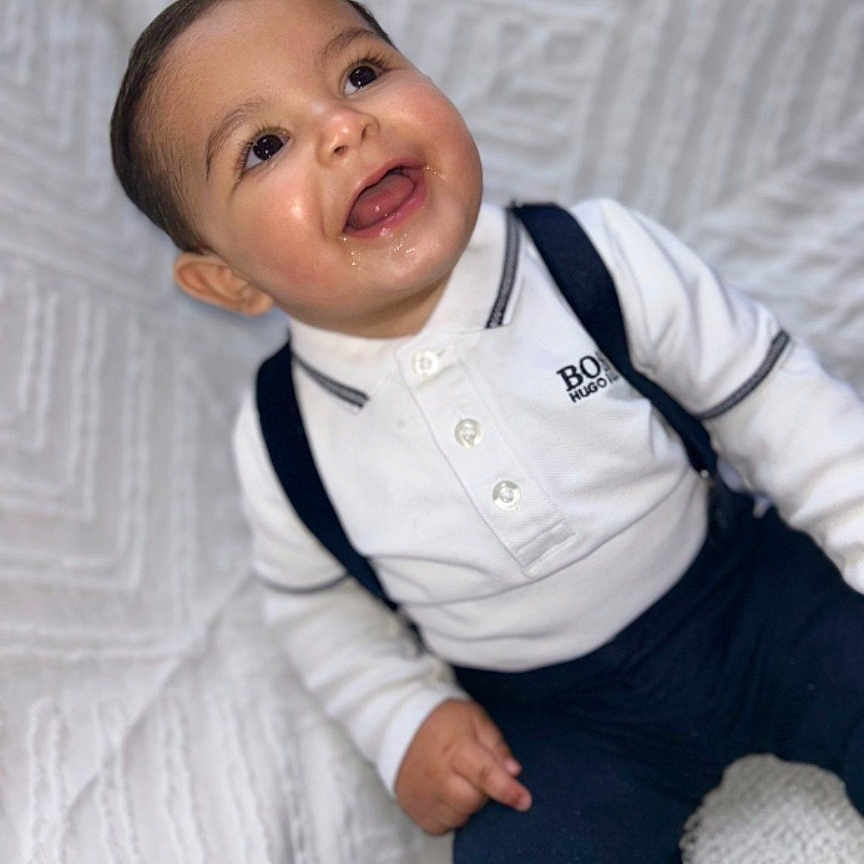Marwan a rejoint le concours — aidez-le/la à gagner de superbes lots ! baby, child, smiling, white_shirt, suspenders, navy_pants, cute, portrait, happy, indoors, texture, blanket, sitting, face, person, infant, young_child, clothing, joyful, expression