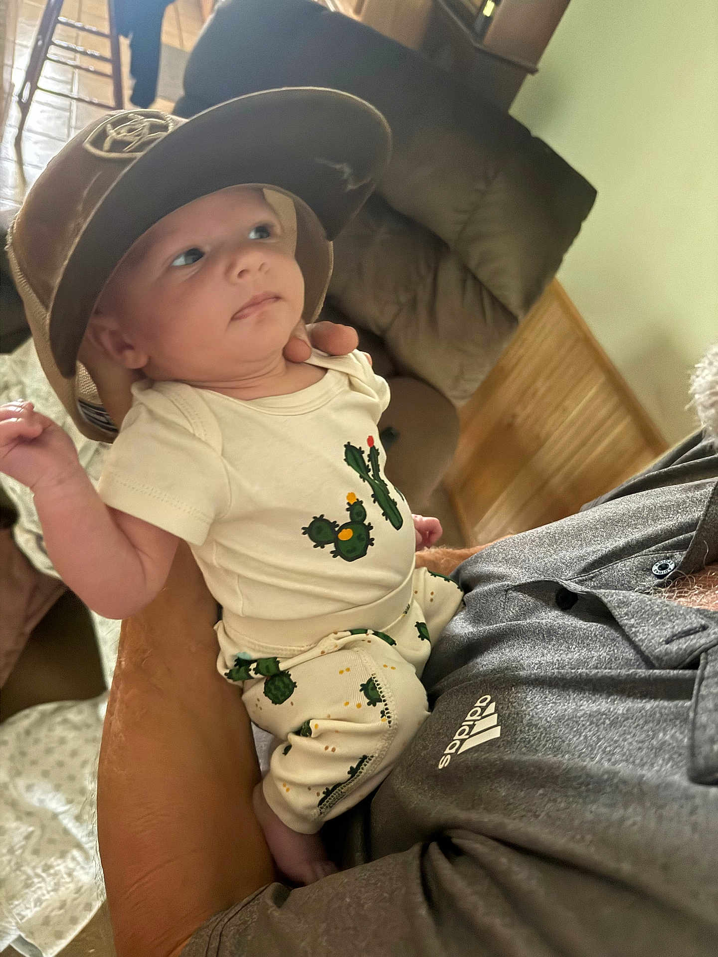 Ryker joined the competition — help win amazing prizes! baby, child, hat, cactus_print, clothing, indoor, person, holding, adult, arm, face, curious, seated, shirt, adidas, couch, furniture, wood_paneling, cozy, home