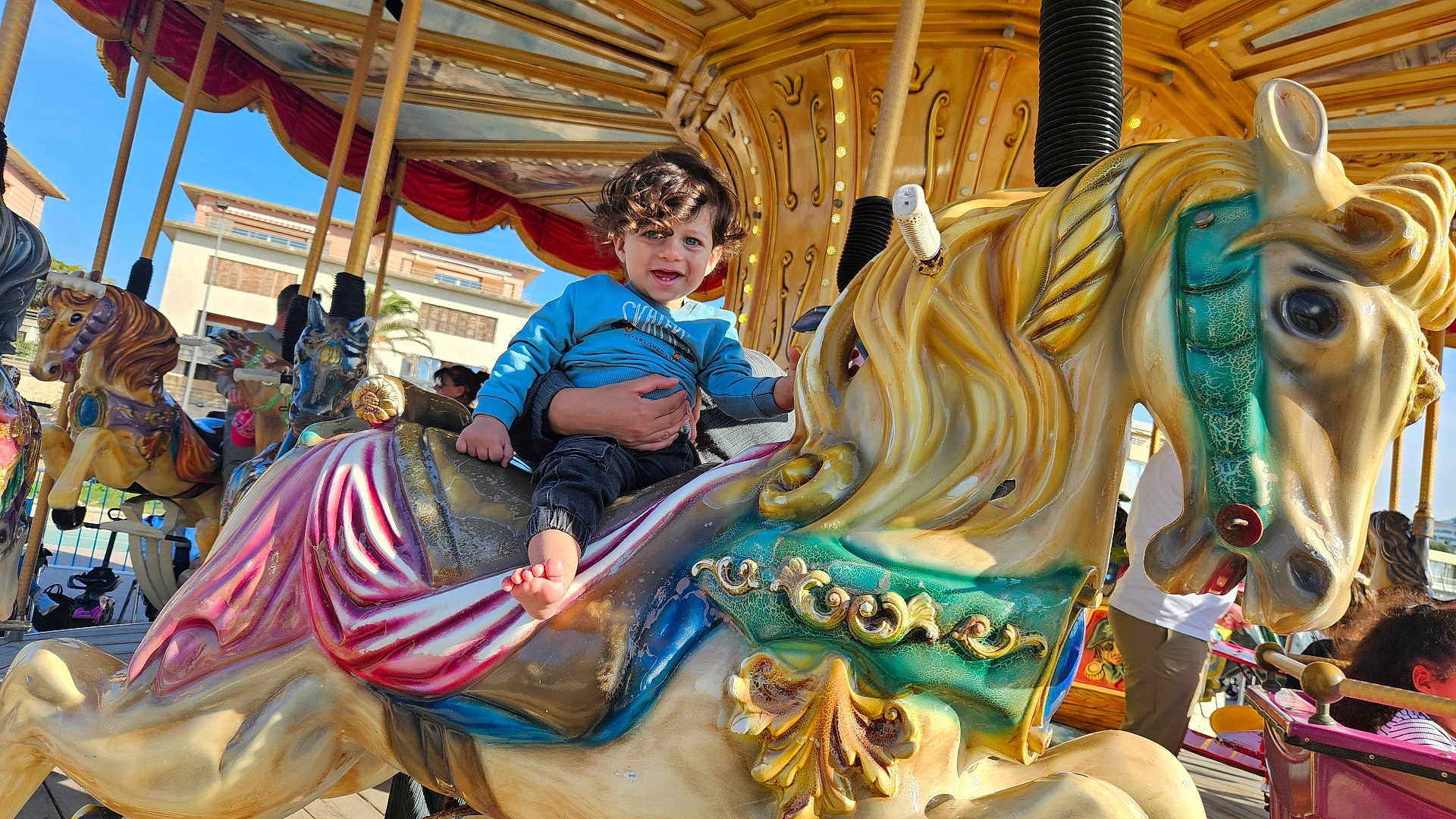 Kenzi a rejoint le concours — aidez-le/la à gagner de superbes lots ! toddler, child, carousel, horse, amusement_park, ride, blue_sky, outdoor, curly_hair, smiling, adult_hand, colorful, toy, play, fun, sunny, daytime, people, background, decorative