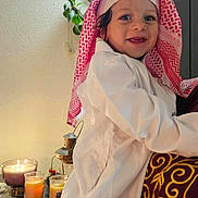 Kenzi participe au concours pour gagner de l'argent avec cette photo : toddler, child, smiling, traditional_clothing, headscarf, white_robe, person, indoor, candles, plant, decor, warm_lighting, holding, adult, maroon_garment, golden_embroidery, happy, face, portrait, cozy