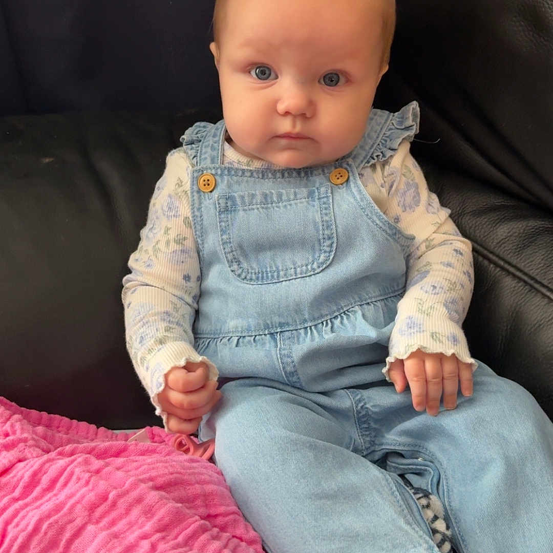 René-Rose joined the competition — help win amazing prizes! baby, blanket, blue_eyes, casual, child, clothing, couch, denim_overalls, expression, face, foot, hand, indoor, infant, long_sleeve_shirt, patterned_blanket, pink_blanket, portrait, sitting, young_child