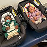 babies, infants, sleeping, car_seats, indoors, portrait, pacifier, pink_outfit, green_outfit, floral_padding, blanket, buckled, transportation, nursery, safety_labels, seated, peaceful, siblings, photograph, cute