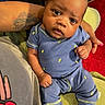 baby, infant, child, portrait, sitting, blue_outfit, onesie, lightning_bolts, wide_eyed, face, smiling, indoor, adult_arm, tattoo, hand, red_background, green_blanket, clothing, close_up, family