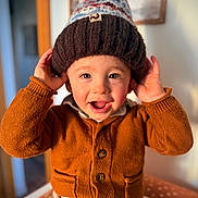Noé participe au concours pour gagner de l'argent avec cette photo : toddler, child, smiling, hat, knitted_hat, pom_pom, orange_cardigan, buttoned_cardigan, hands_up, happy, indoor, portrait, face, cute, warm_clothing, cozy, baby, light, blurred_background, person