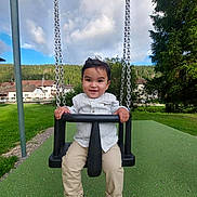 Lorenzo a rejoint le concours — aidez-le/la à gagner de superbes lots ! child, toddler, swing, playground, outdoor, grass, trees, sky, clouds, houses, greenery, sandals, pants, shirt, happy, smile, person, daylight, nature, fun